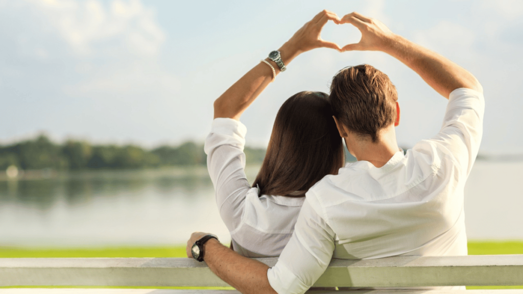 Casal sentado em um banco ao ar livre, formando um coração com as mãos, enquanto apreciam a vista de um lago sereno e arborizado ao fundo.