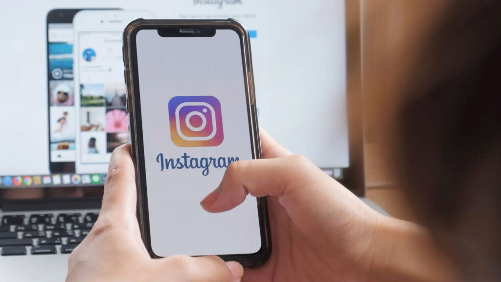 Uma pessoa segurando um smartphone que exibe a tela inicial do Instagram, com o logo característico em grande destaque. No fundo, um computador com várias telas abertas também mostra conteúdo relacionado ao Instagram, indicando um ambiente de trabalho ou de gestão de mídia social.