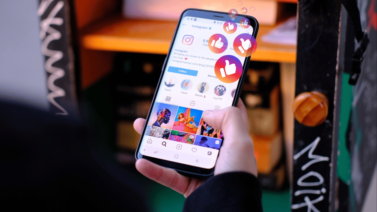 Uma pessoa segurando um smartphone com a tela do Instagram aberta, mostrando o feed com várias postagens coloridas. A imagem é capturada em um ambiente interno, com foco na tela do telefone que exibe ícones de curtidas e notificações flutuantes em um design atraente e moderno.