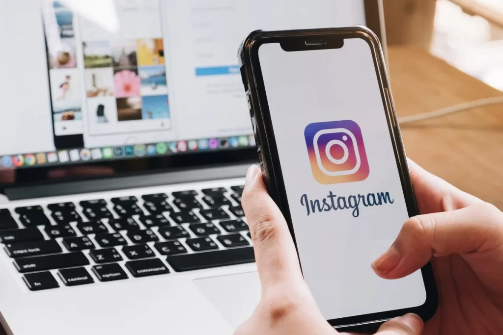 Uma pessoa segurando um smartphone com o logotipo do Instagram exibido na tela, em foco. No fundo desfocado, um laptop com o teclado visível, indicando que também está sendo usado para acessar o Instagram ou outras tarefas relacionadas.